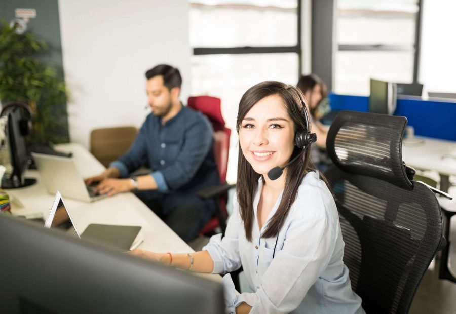 Portrait of pretty young latin woman wearing a headset sitting at her office desk with coworkers in background