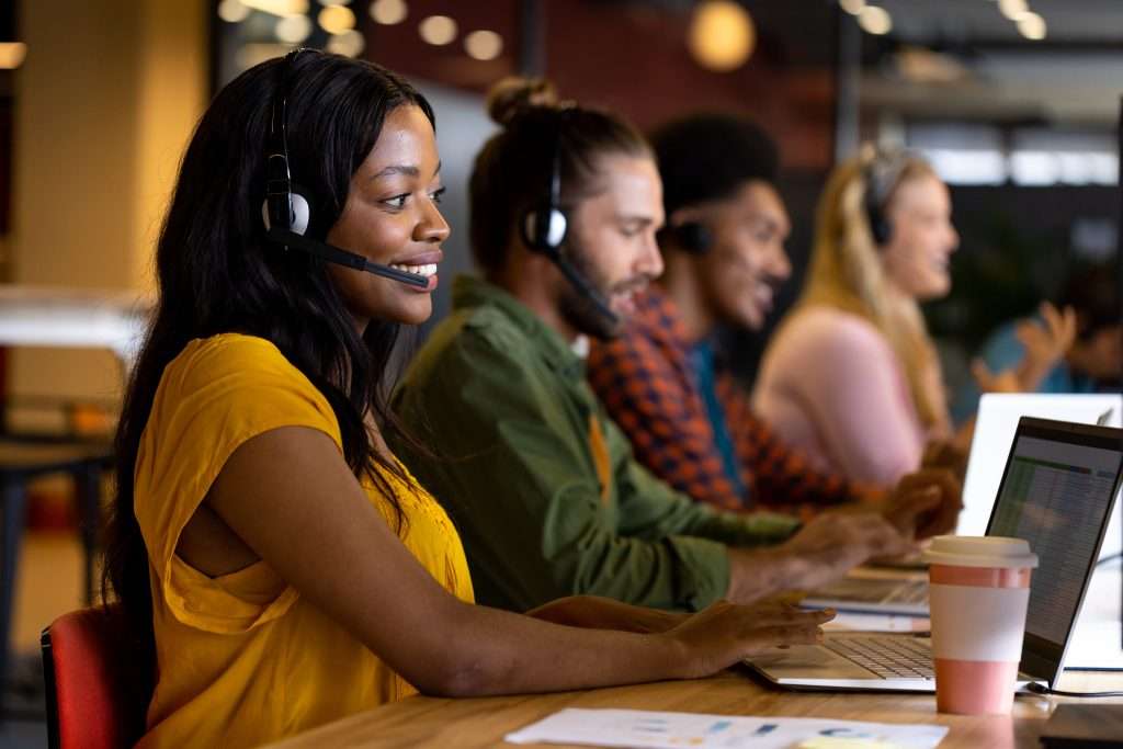 Happy diverse colleagues in phone headsets using computers in office. Casual office, business, communication, teamwork and work, unaltered. Diverse call center team
